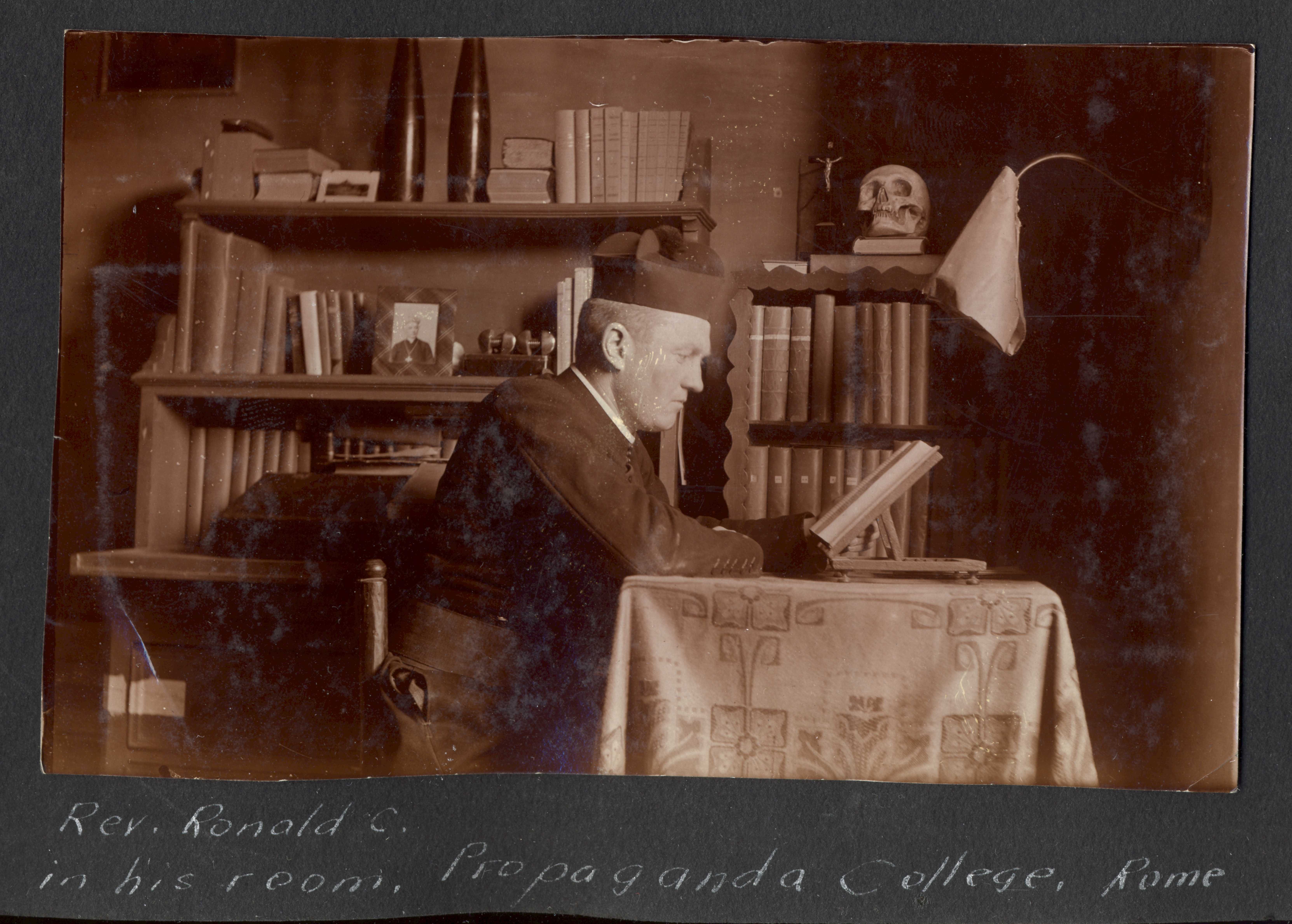 Black and white photograph. RC MacGillivray is shown from the side, sitting at a table covered in a long, embroidered cloth, reading from a book. There are book shelves behind him. He is wearing a dark uniform and a hat.